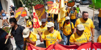 Photo of a group of people marching down a street wearing bright yellow t-shirts reading &quot;JUSTICE for Farmworkers&quot; and carrying colorful banners and signs featuring tomatoes that read &quot;no more exploitation&quot;  and &quot;no more abuses.&quot; 