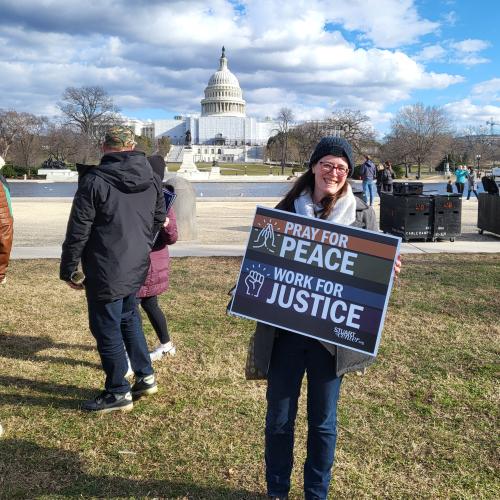 A white woman, bundled against the cold, stands before the US Capitol with a sign reading &quot;Pray for Peace, Work for Justice&quot;