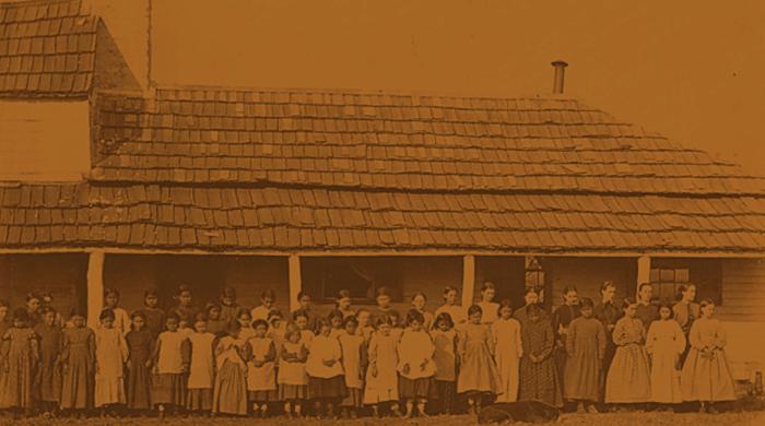 Sepia tinted photograph of a large group of Native American children posed standing in front of a school.