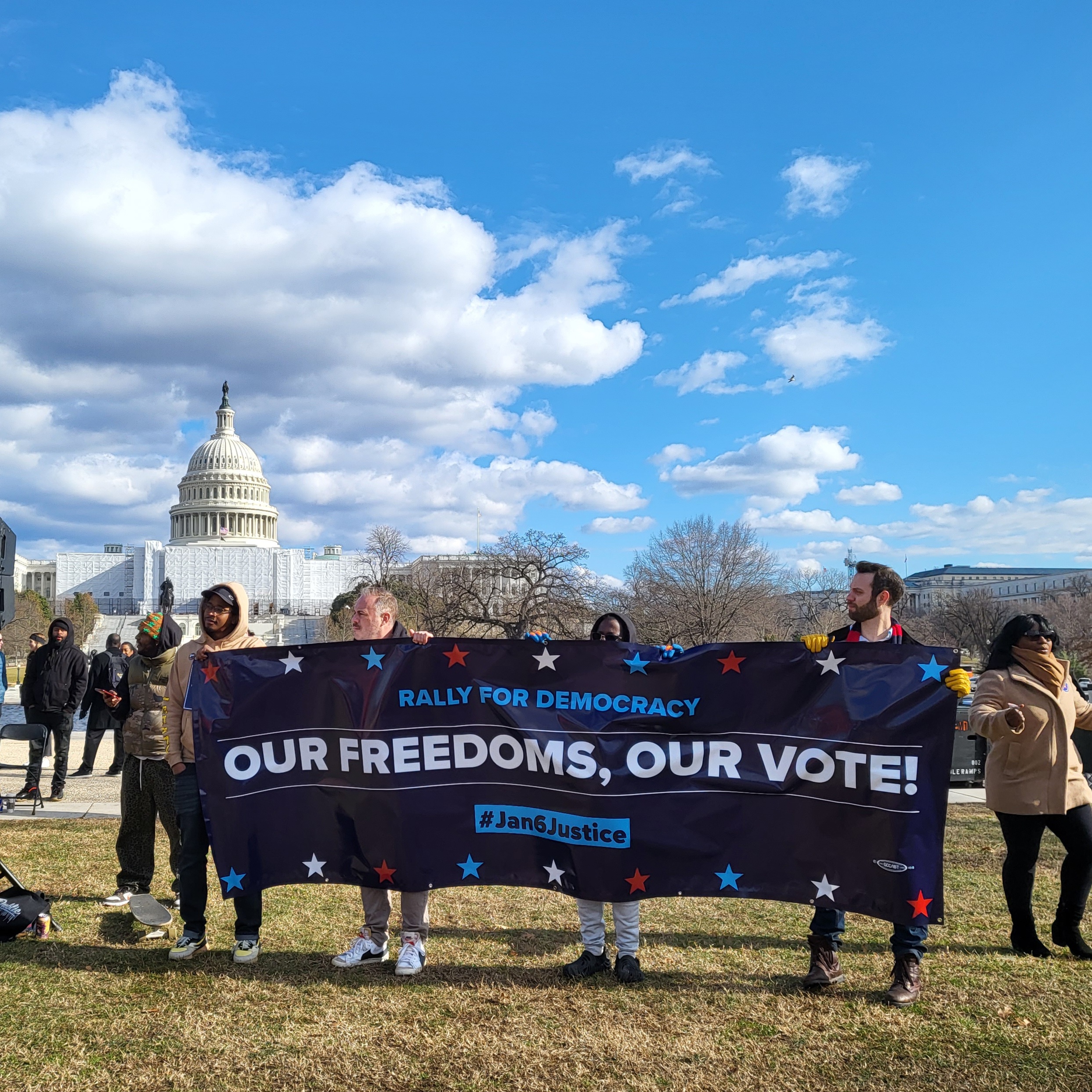 Four Black and white men stand holding a banner reading "Rally for Democracy; Our Freedoms, Our Vote - #Jan6Justice"